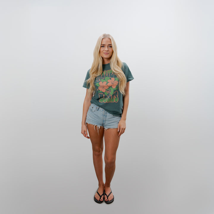 Young woman wearing a green Malibu Aloha Tee with floral graphics, paired with light denim shorts and flip-flops, standing against a plain white background.