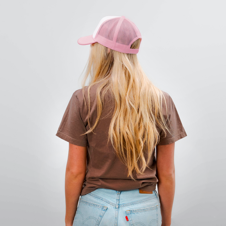 Back view of a woman wearing a pink mesh trucker hat, brown t-shirt, and light blue denim shorts against a plain background.