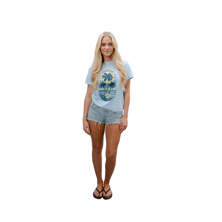 Young woman wearing a light blue Malibu Beach T-shirt with palm trees and ocean graphic, paired with denim shorts and flip-flops.