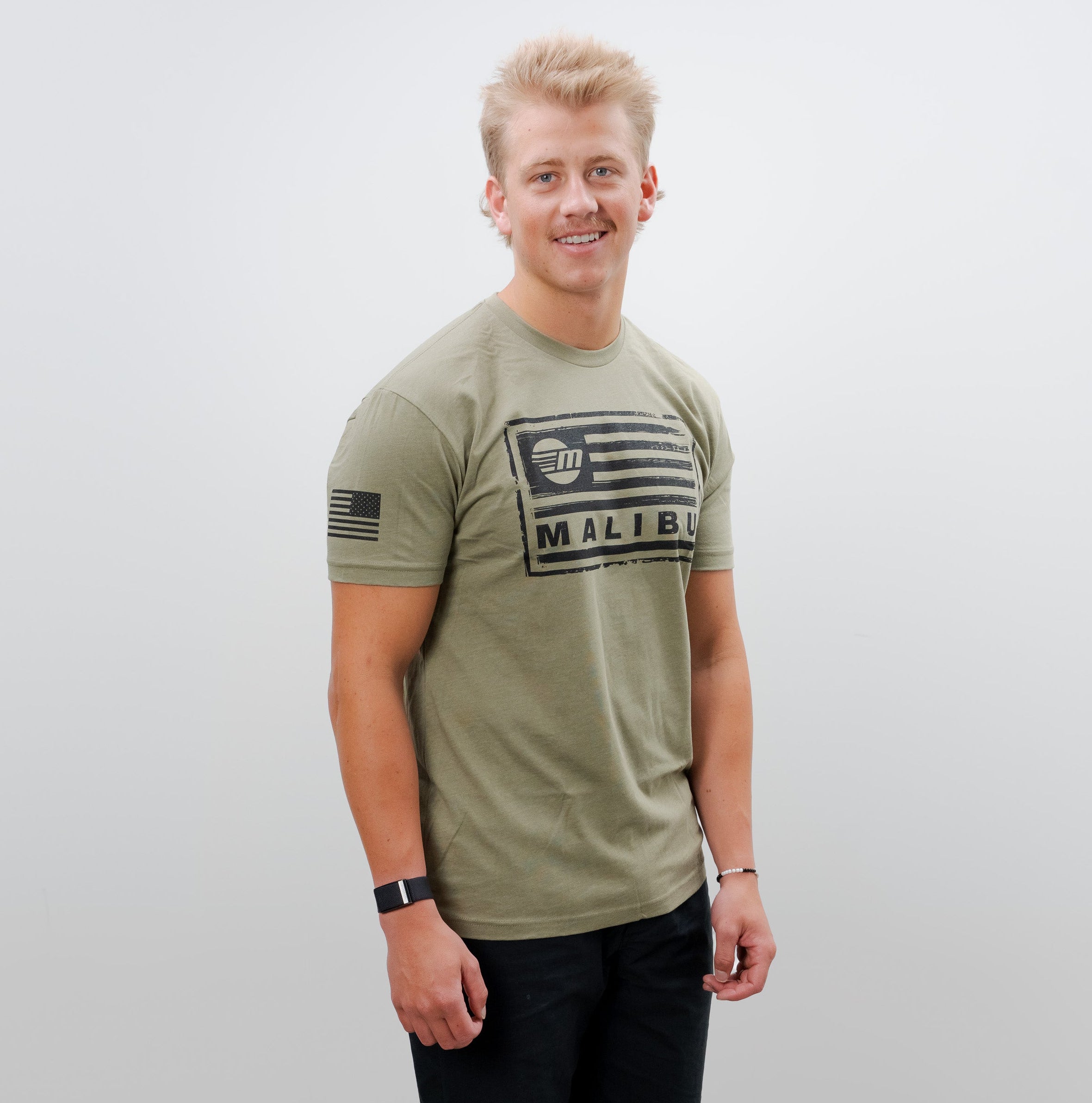 Young man wearing a light green Malibu Freedom Tee 2.0 with black graphic print, black pants, and sneakers, standing against a plain white background.