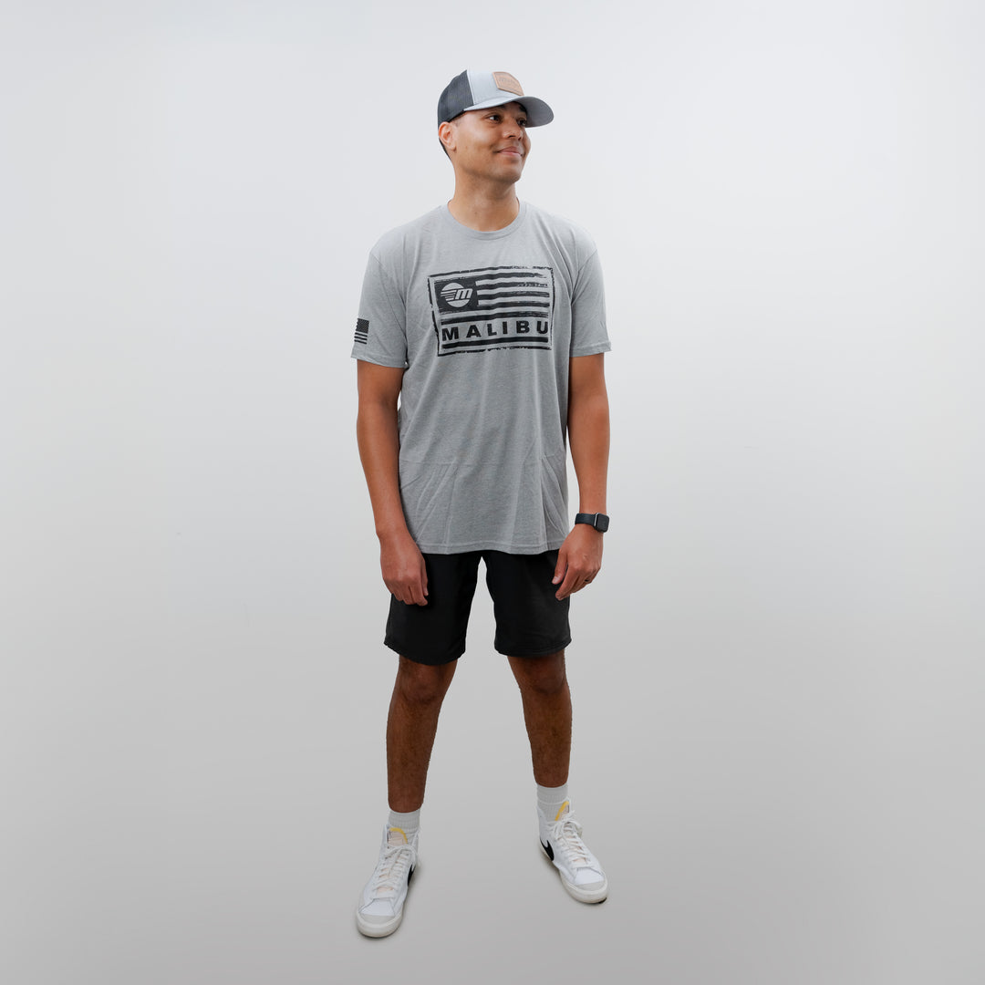 Man wearing a gray Malibu Freedom Tee 2.0 with black shorts, white sneakers, gray cap, and a watch, standing against a plain white background.