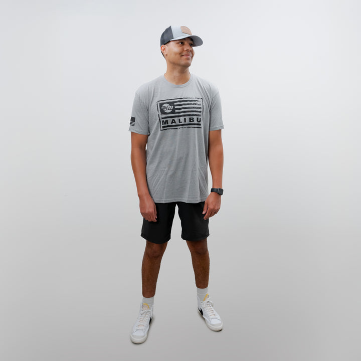 Man wearing a gray Malibu Freedom Tee 2.0 with black shorts, white sneakers, gray cap, and a watch, standing against a plain white background.