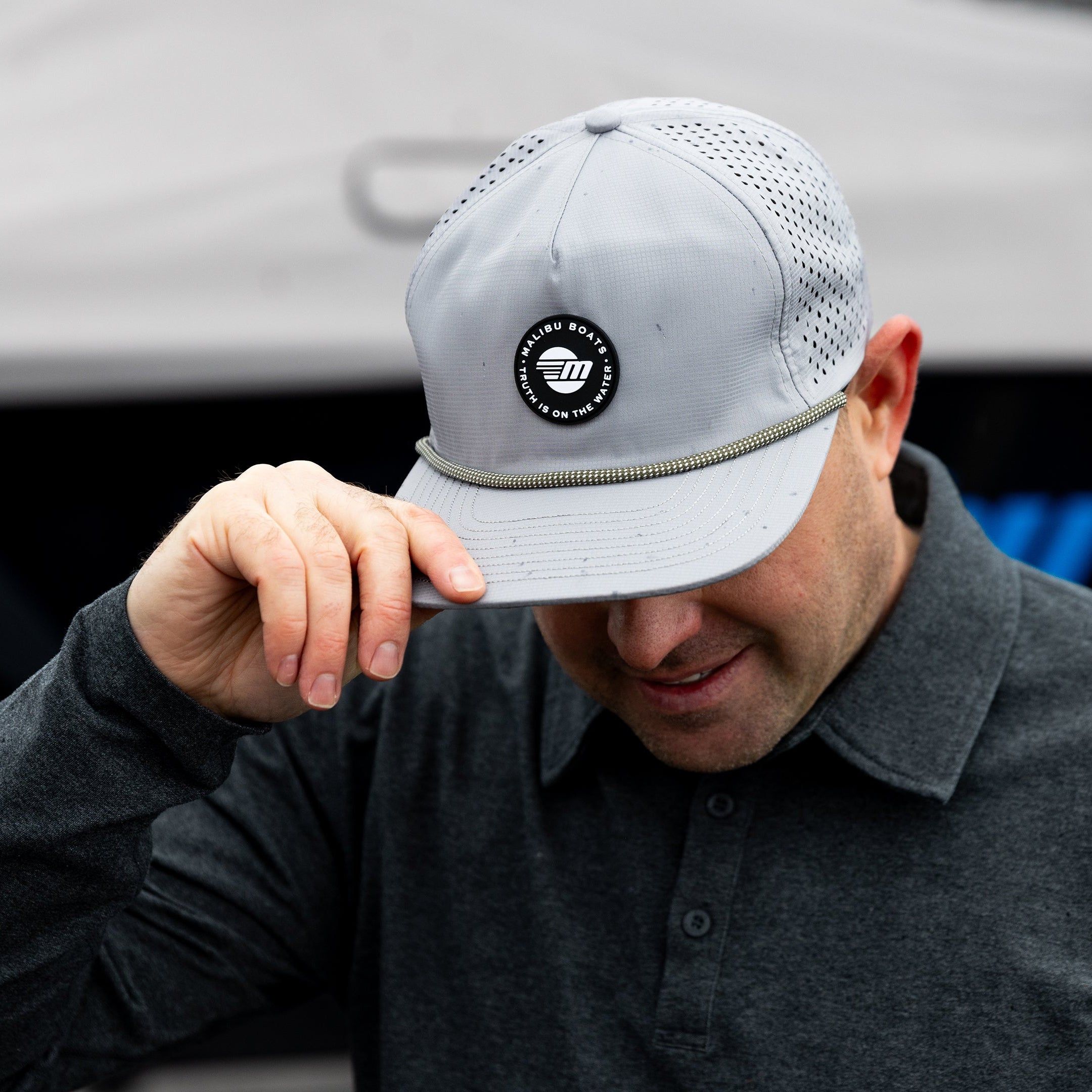 Man wearing a light gray Malibu Phantom hat with a circular logo, adjusting the brim while outdoors near a boat.
