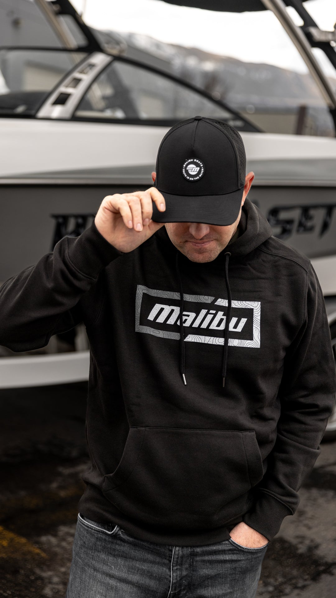 Man wearing a black Malibu Truth Hat, hoodie, and jeans, standing in front of a boat.
