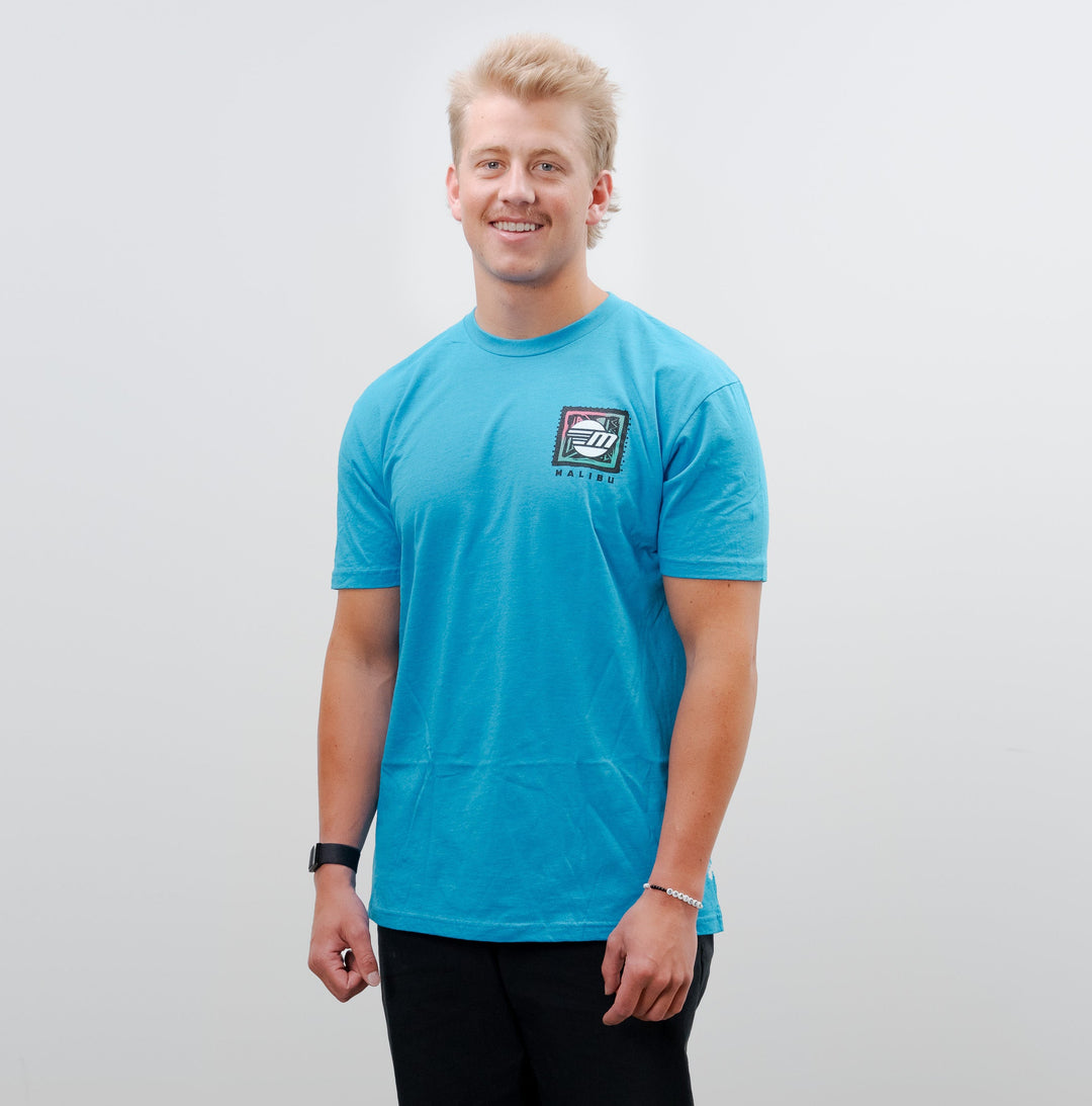 Young man smiling in a blue Malibu Wave Warrior t-shirt, black pants, and sneakers, standing against a plain white background.