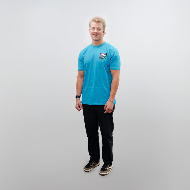 A young man stands wearing a blue Malibu Wave Warrior t-shirt, paired with black pants and sneakers, showcasing a casual style.