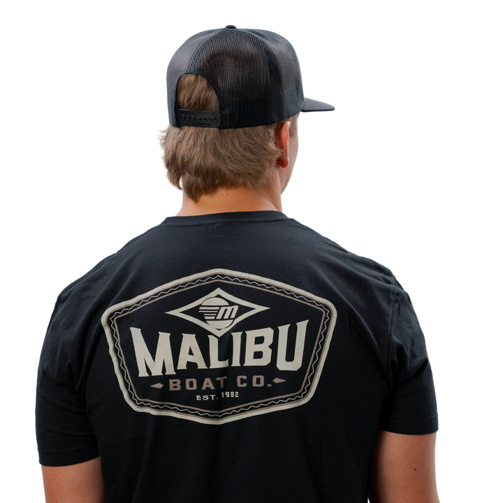 Back view of a person wearing a black Malibu Diamond Tee with a large Malibu Boat Co. logo, and a black mesh trucker cap.