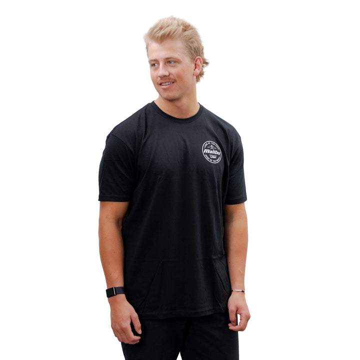 Male model wearing a black Malibu Brave T-shirt with a small logo on the chest, paired with black pants and sneakers, standing against a plain white background.