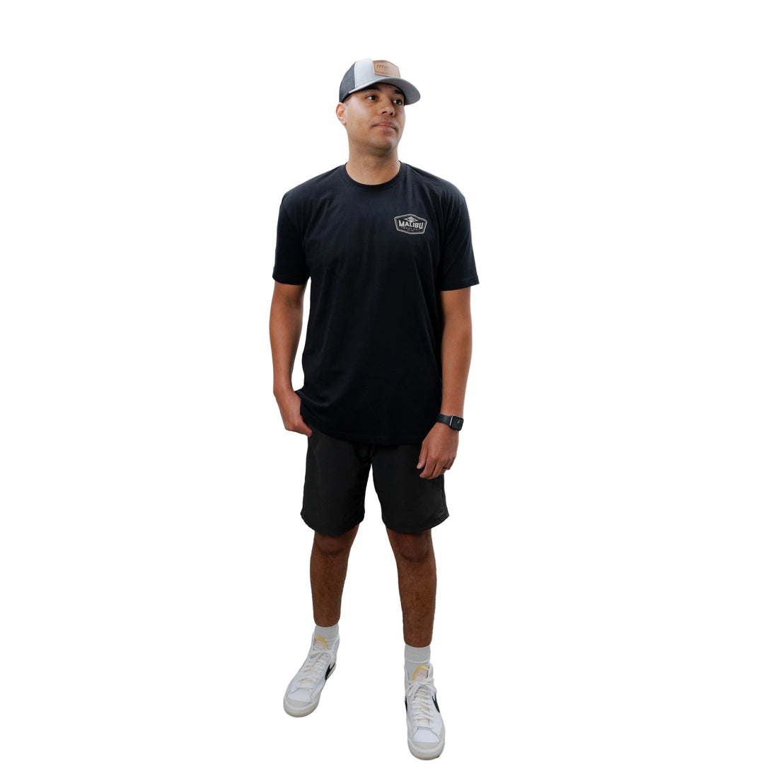Man wearing a black Malibu Diamond Tee, black shorts, white sneakers, and a gray cap, standing against a plain white background.