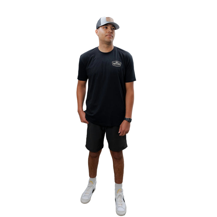 Man wearing a black Malibu Diamond Tee, black shorts, white sneakers, and a gray cap, standing against a plain white background.