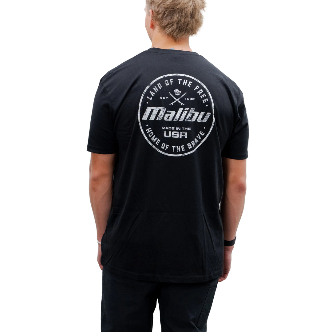 Back view of a person wearing a black Malibu Brave T-shirt with a circular logo design on the back featuring text and graphics.