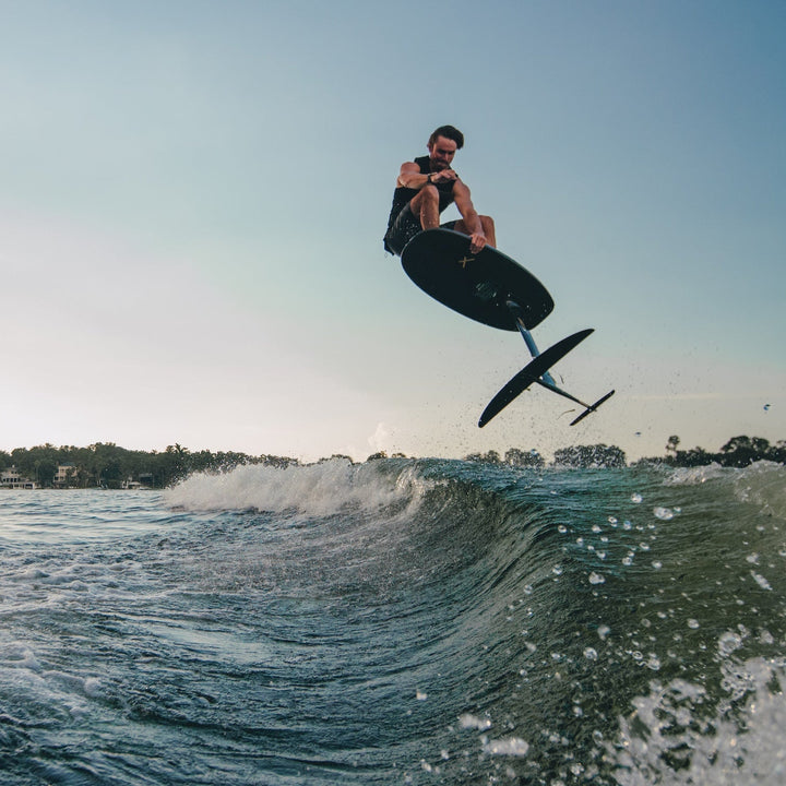 A person performs a jump on a surfboard with a hydrofoil over ocean waves during sunset.