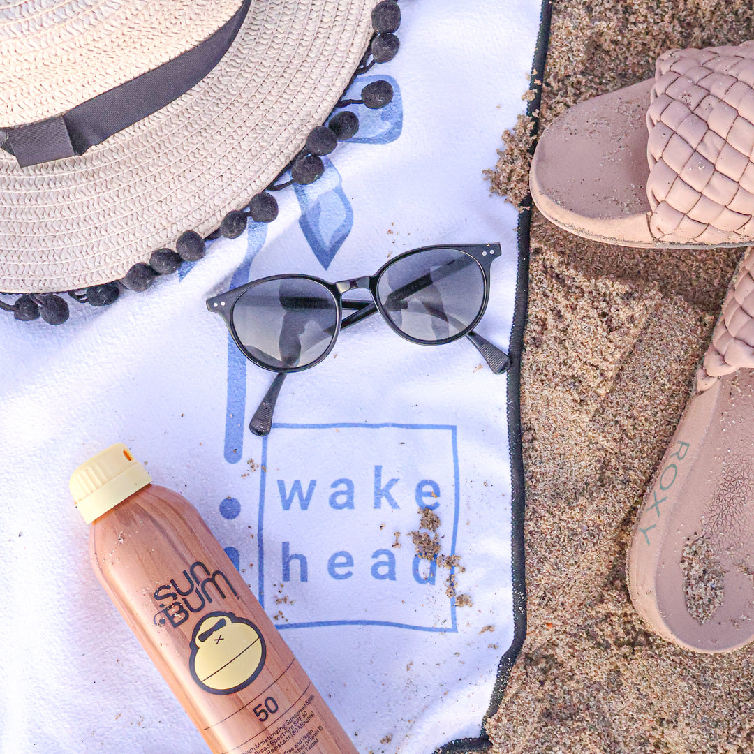 Blue Bliss Lake Towel with "wake head" logo, surrounded by sunglasses, a straw hat, a sunscreen bottle, and sand on a beach.