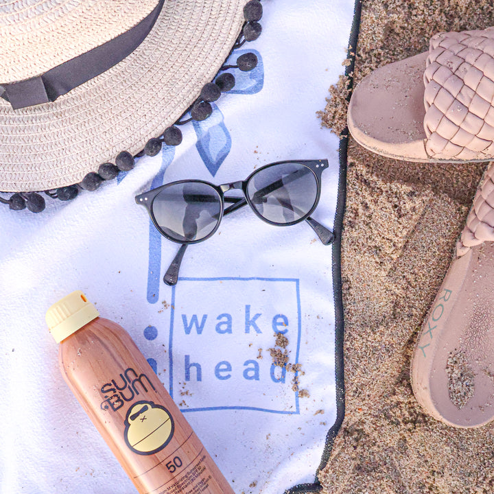 Blue Bliss Lake Towel with "wake head" logo, surrounded by sunglasses, a straw hat, a sunscreen bottle, and sand on a beach.