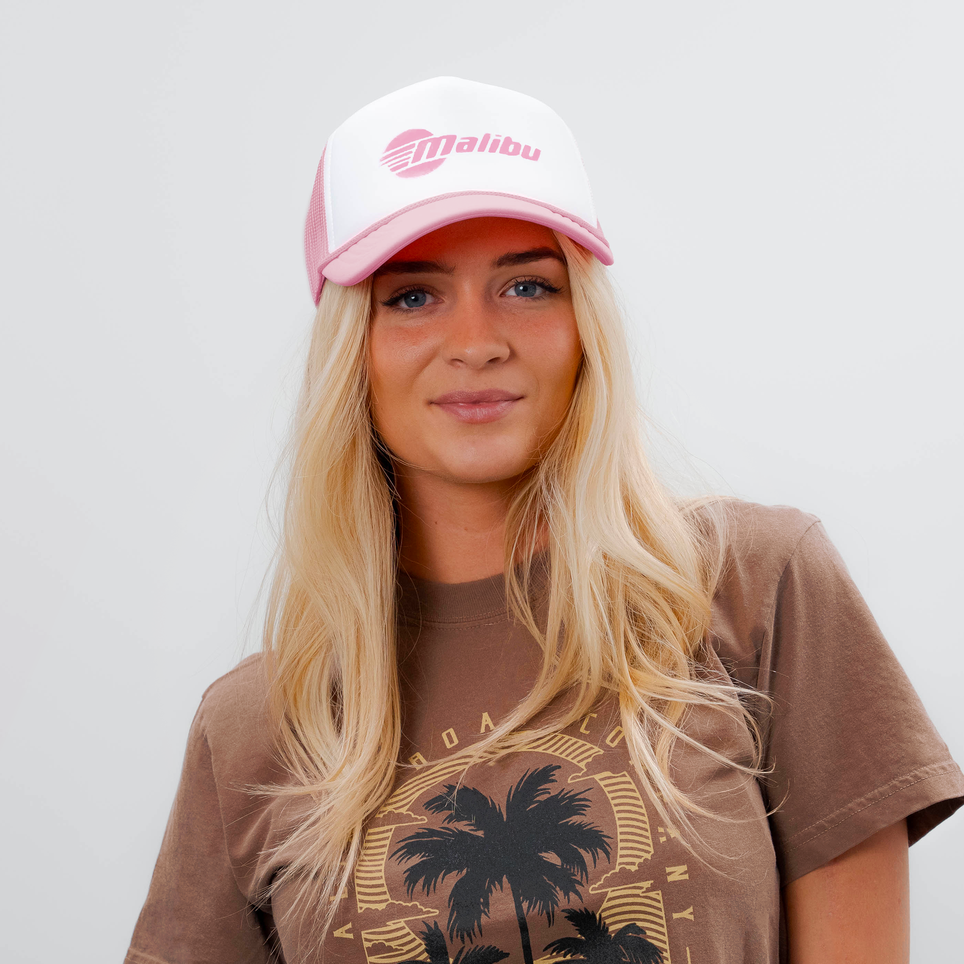A woman wears a pink and white Malibu trucker hat with a mesh back, featuring a pink Malibu logo on the front.