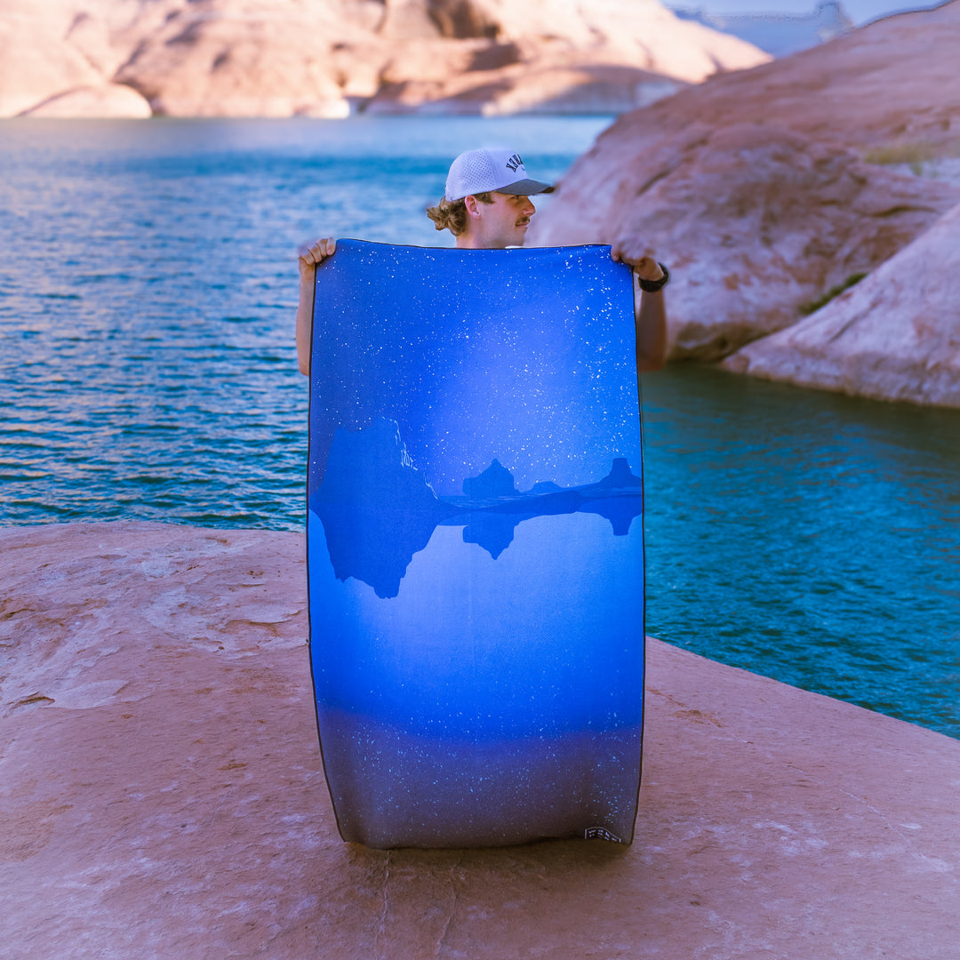 A person holds a Canyon Dreams wake head towel featuring a blue starry design by a lake, surrounded by rocky terrain.