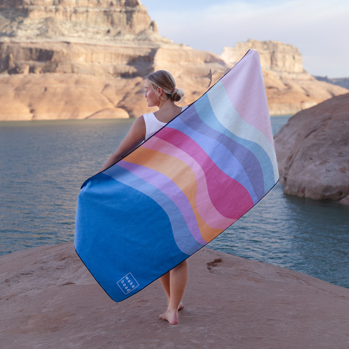 A woman stands on a rocky shore holding a pastel-colored lake towel with wavy patterns, overlooking a serene lake.