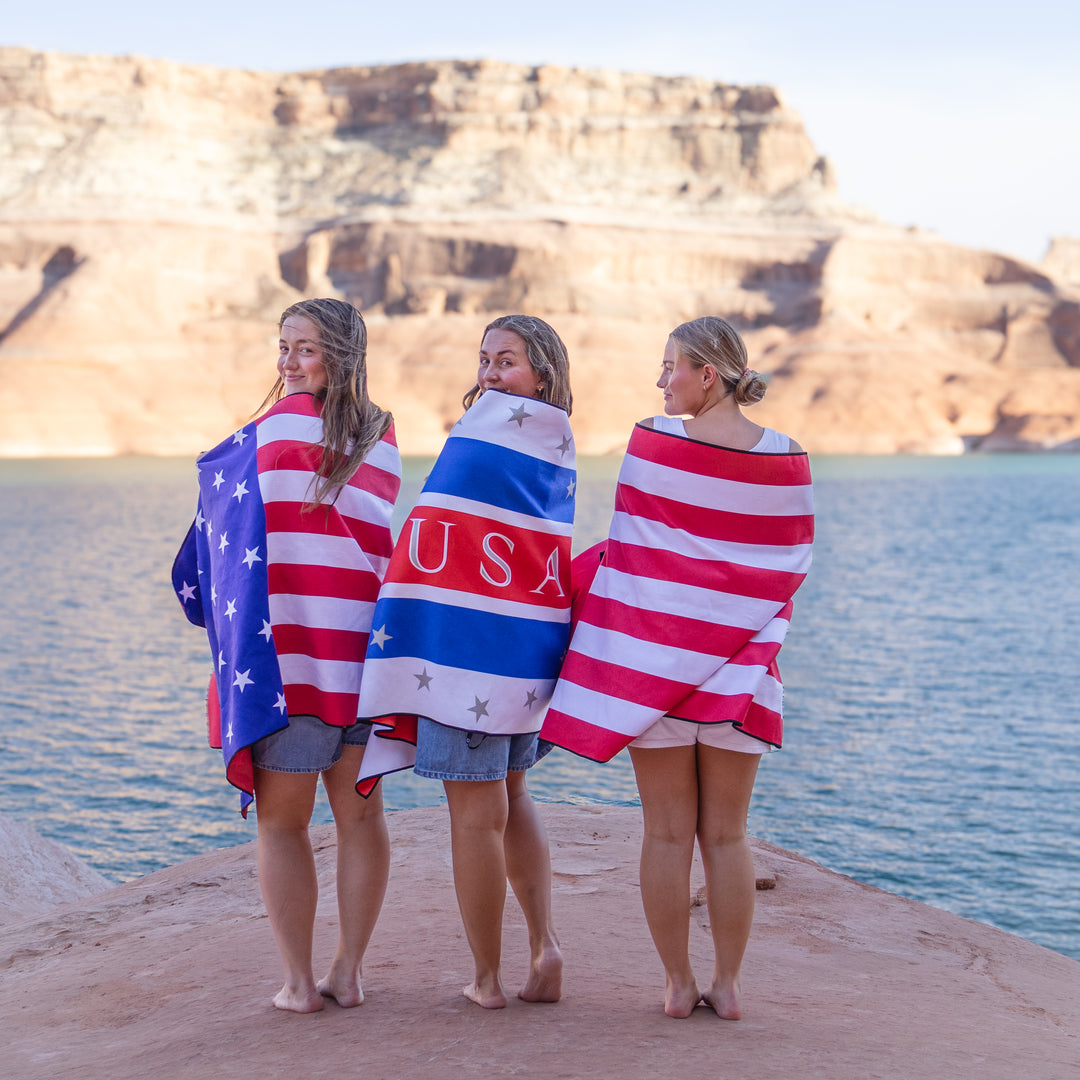 Three women wrapped in a Stars &amp; Stripes lake towel stand by the water, showcasing a patriotic design against a scenic backdrop.