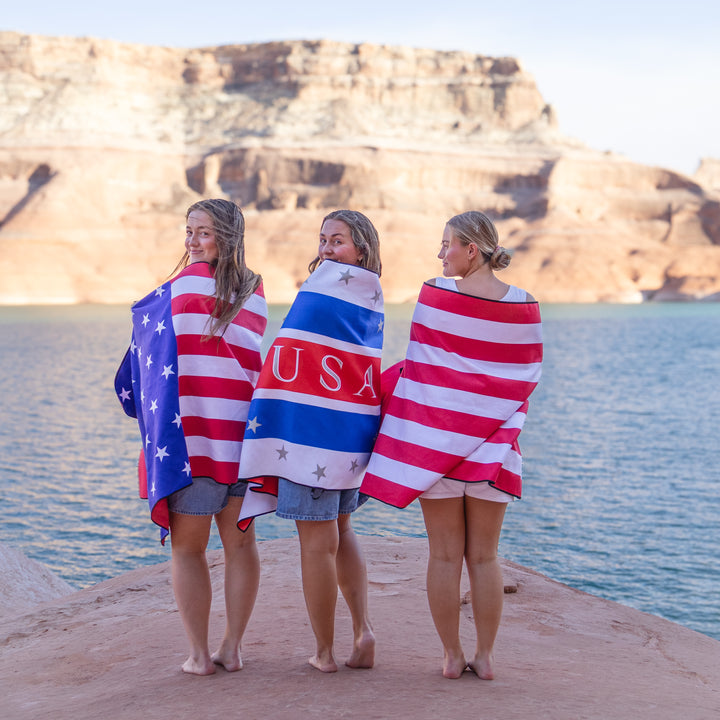 Three women wrapped in a Stars &amp; Stripes lake towel stand by the water, showcasing a patriotic design against a scenic backdrop.