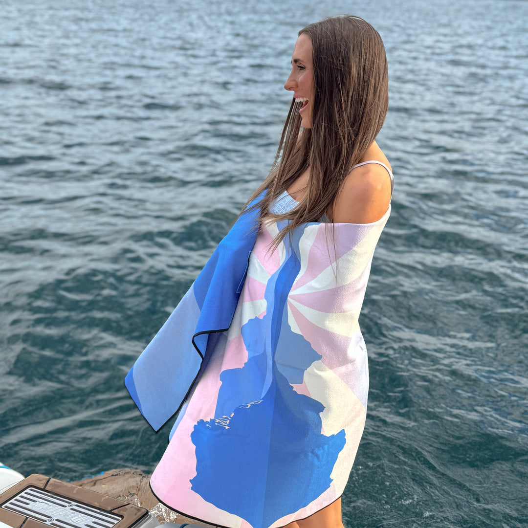 A woman stands by the water wrapped in a colorful Sunset Oasis towel, showcasing its vibrant design and beach vibe.
