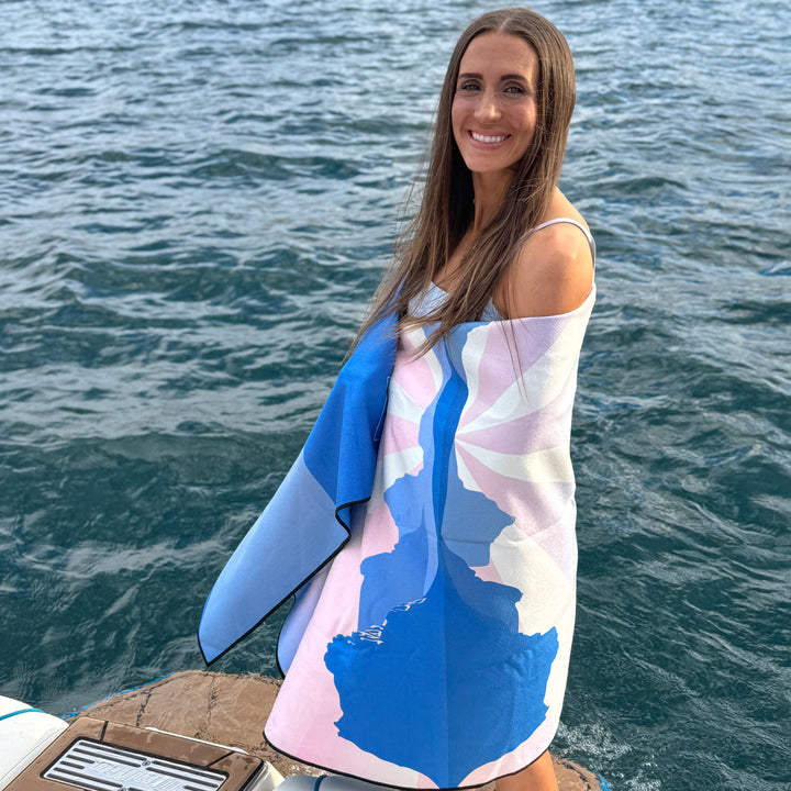 A woman stands on a boat wrapped in a colorful Sunset Oasis towel, with water in the background.
