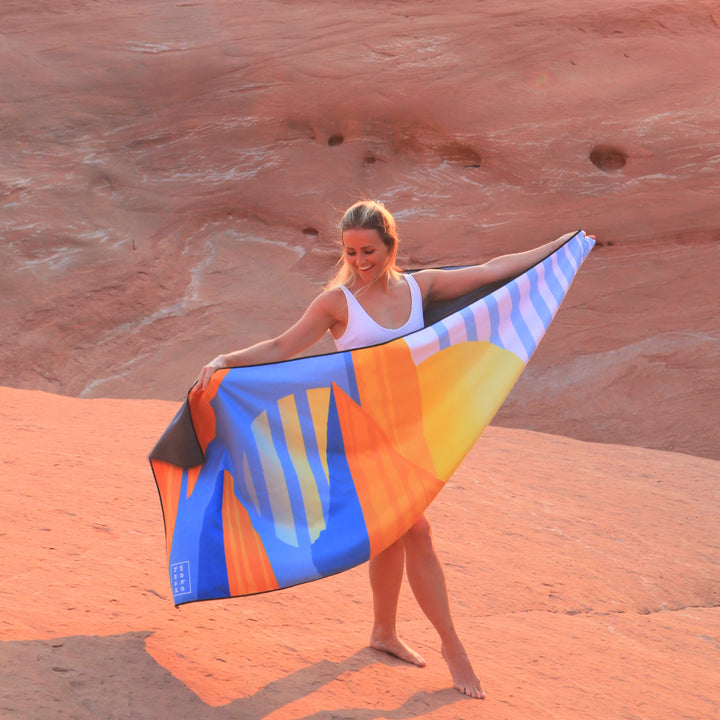 Woman smiling on desert terrain holding a colorful Powell Towel with blue, orange, yellow, and white stripes.