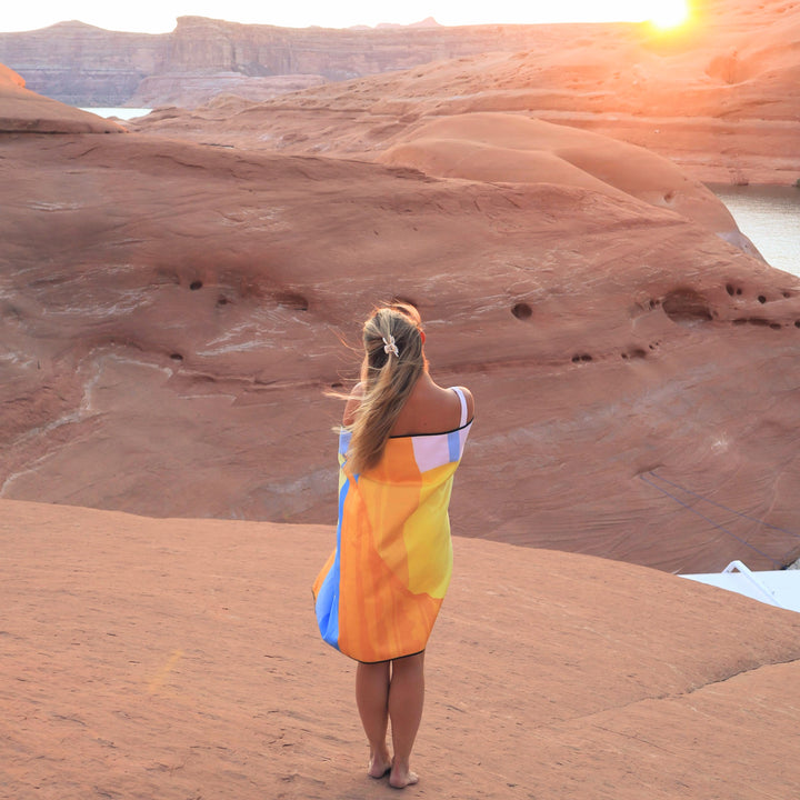 A woman standing on desert rocks wrapped in a colorful Powell Towel, overlooking a scenic canyon at sunset.