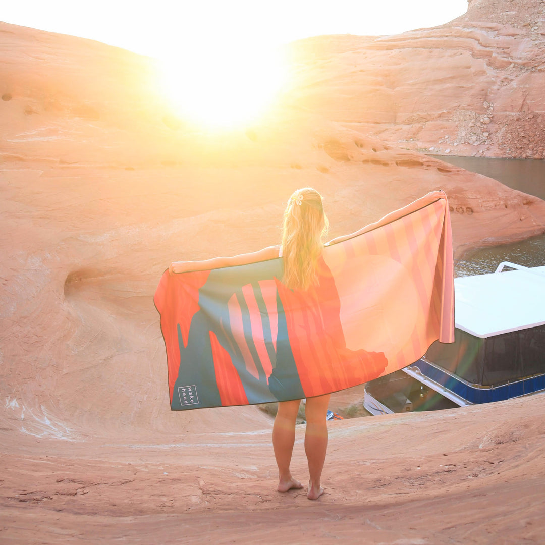 Woman holding a Powell Towel at sunset in a desert canyon, with water and boat nearby, enjoying a relaxing moment.