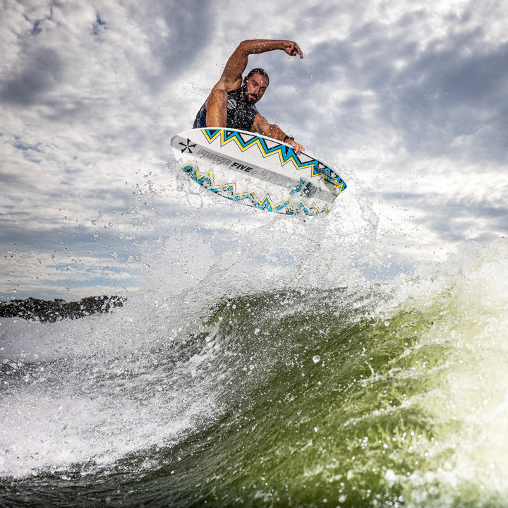 A surfer performs a jump on the 2026 Phase Five Ahi board, creating a splash in the water under a cloudy sky.