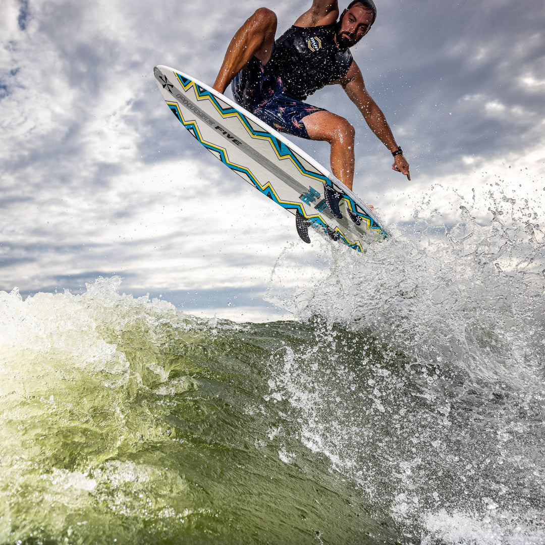 A surfer performs an aerial maneuver on a 2026 Phase Five Ahi board, splashing through ocean waves under a cloudy sky.