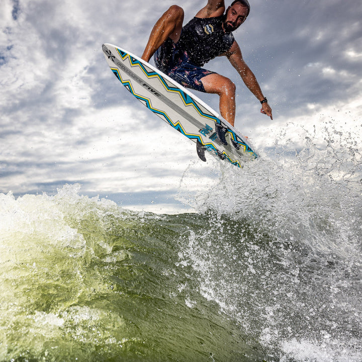 A surfer performs an aerial maneuver on a 2026 Phase Five Ahi board, splashing through ocean waves under a cloudy sky.
