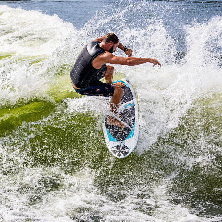 A surfer performs a trick on a 2026 Phase Five Ahi board, riding a wave with splashing water around him.