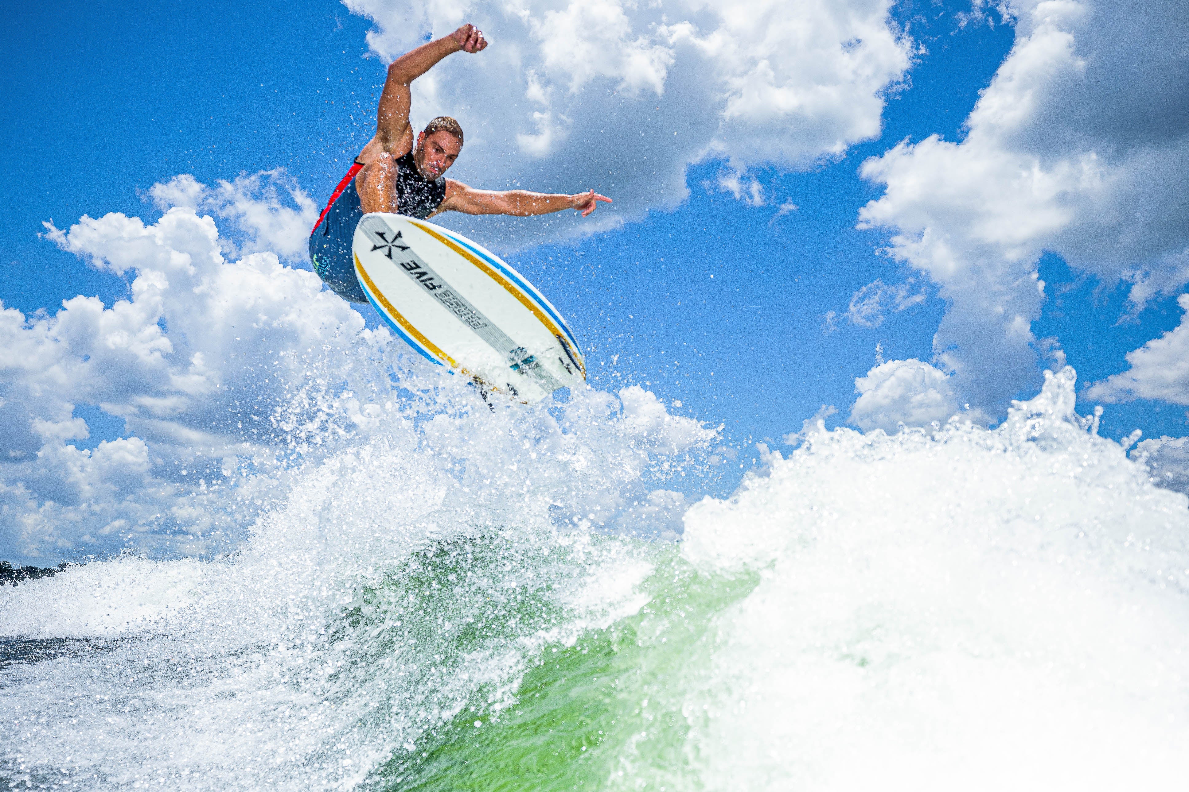 Surfer performing an aerial trick on a 2025 Phase 5 Ahi Surf Board against a bright blue sky with clouds.