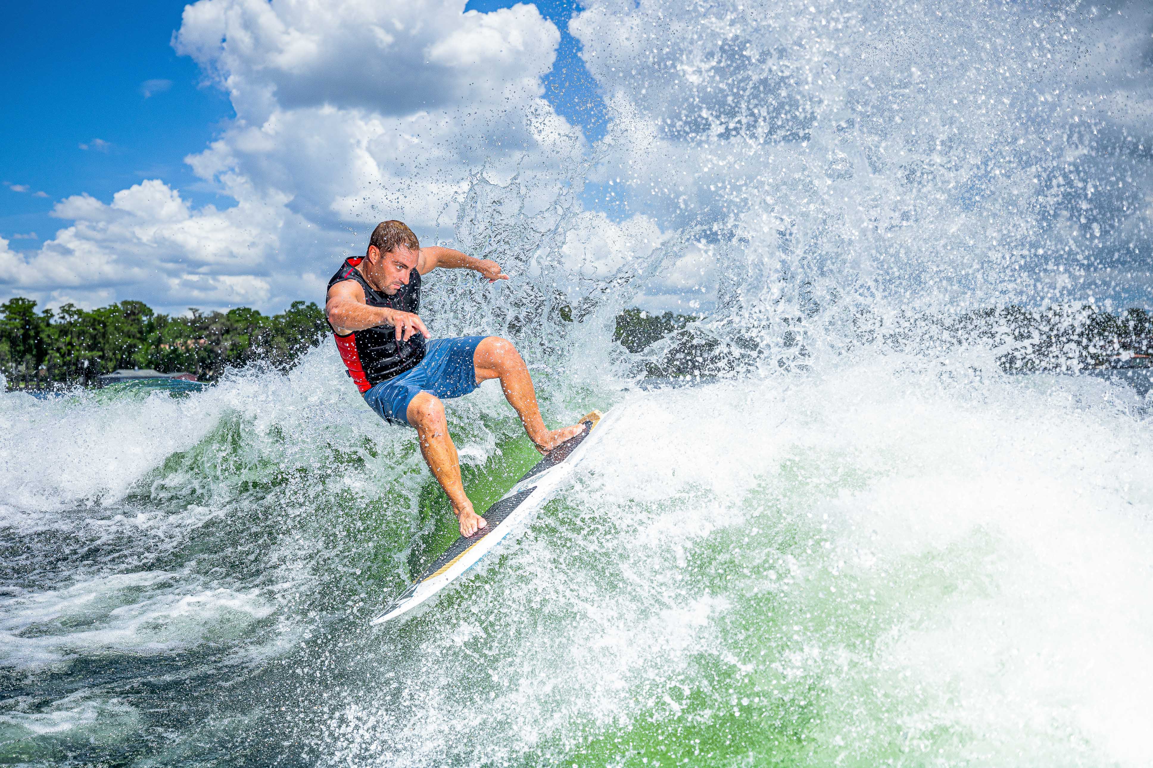 A man surfing on a 2025 Phase 5 Ahi surfboard, riding a wave with splashing water under a partly cloudy sky.