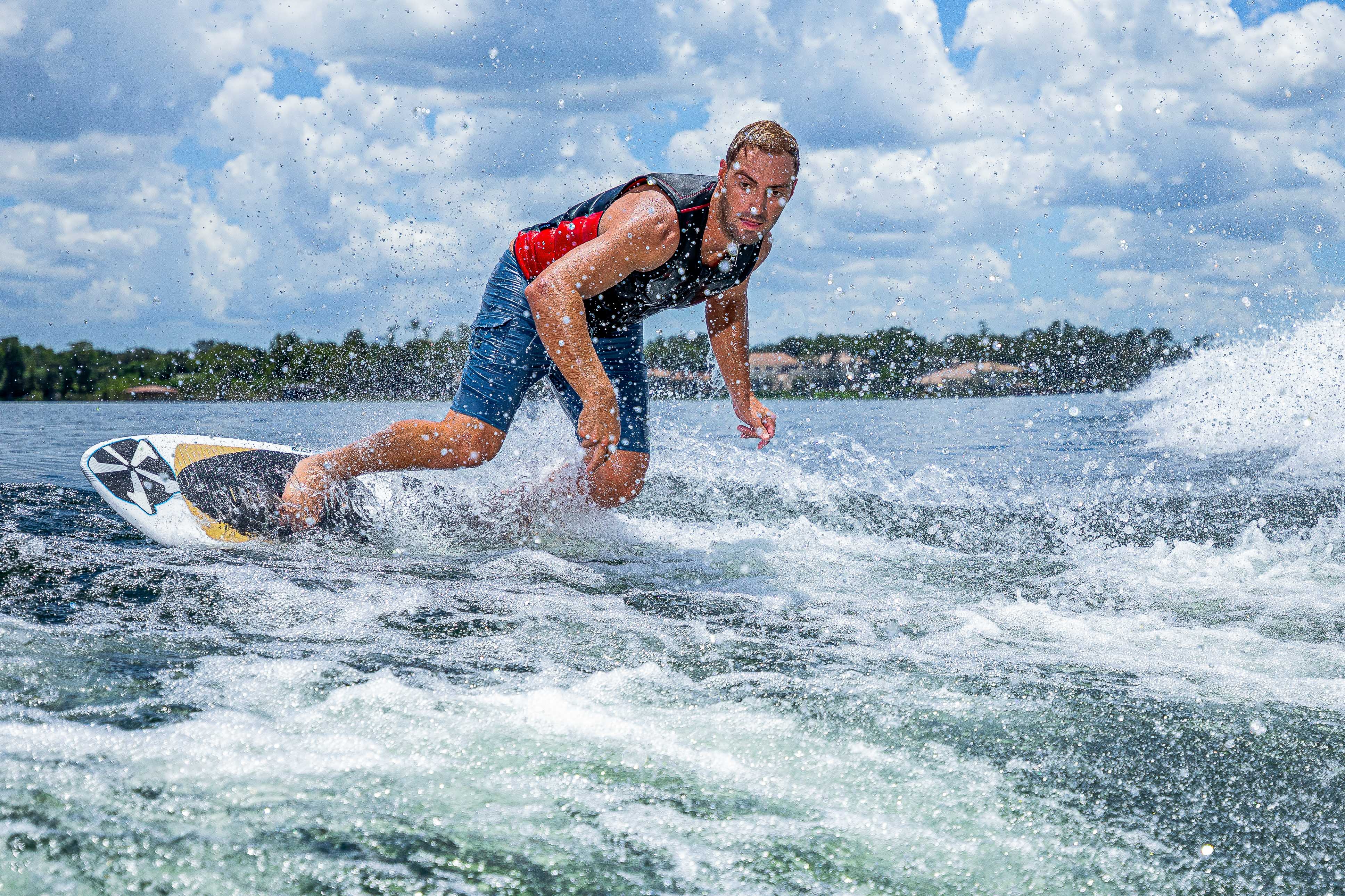 A man surfing on a 2025 Phase 5 Ahi surfboard on a body of water with a cloudy sky and distant shoreline in the background.