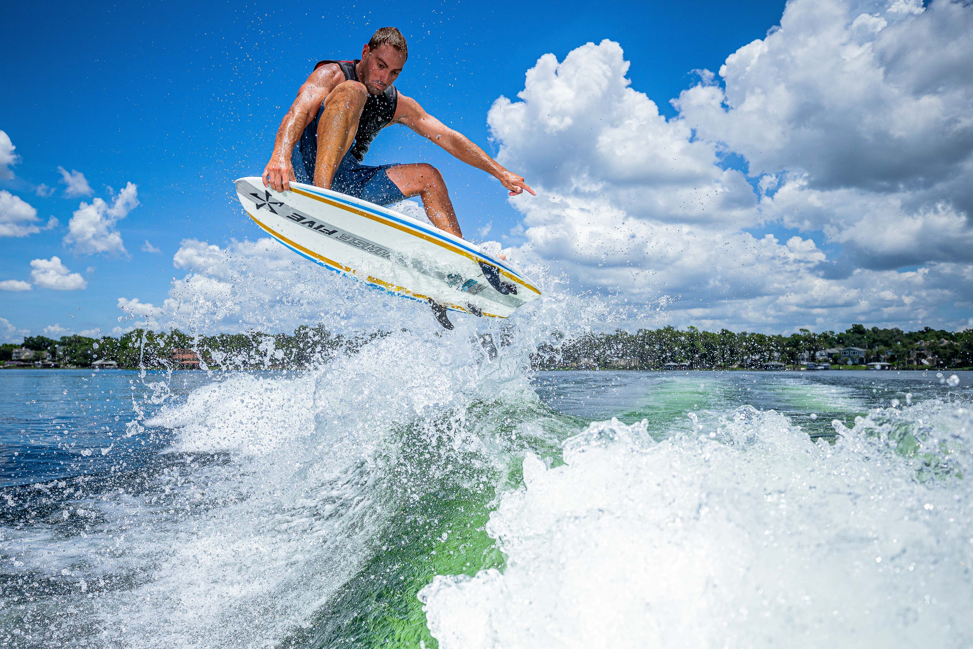 A male surfer performs an aerial trick on the 2025 Phase 5 Ahi Surf Board over a lake with a blue sky and scattered clouds.