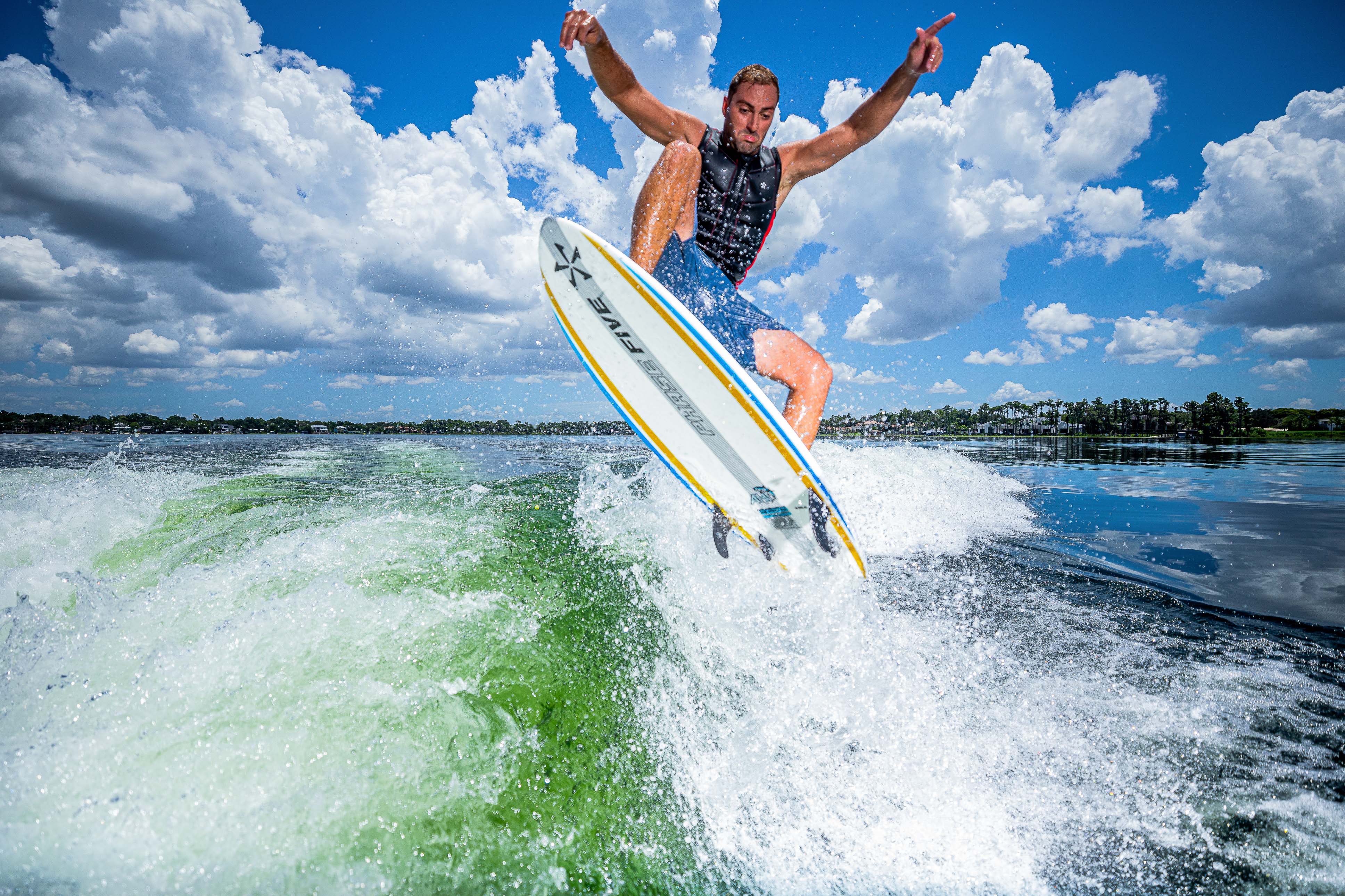A man surfing on a 2025 Phase 5 Ahi surfboard, mid-air above green water with a blue sky and clouds in the background.