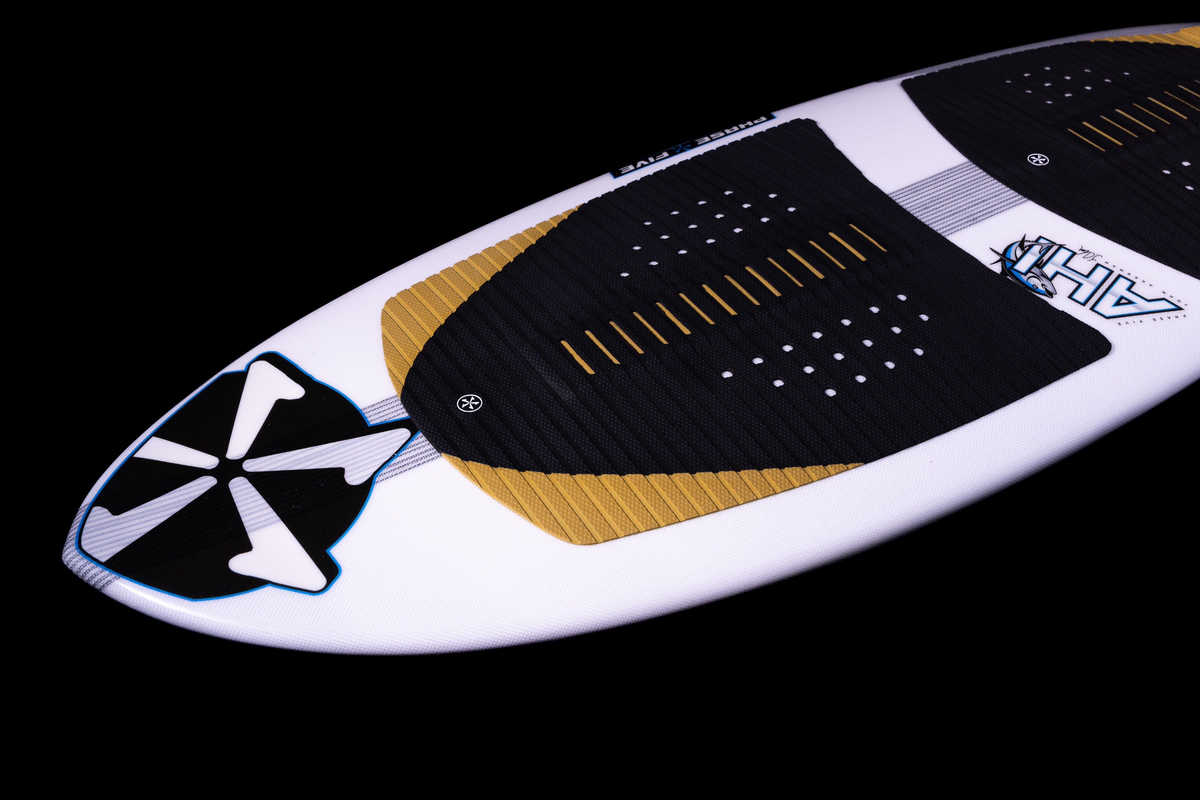 Close-up of the 2025 Phase 5 Ahi surfboard with black traction pads, white, gold, and blue accents on a black background.