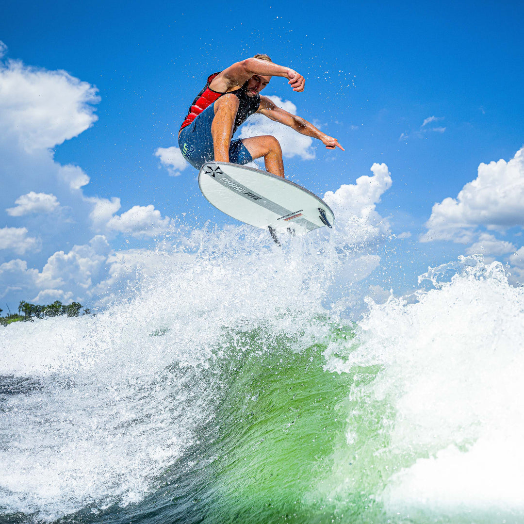 A surfer performs a jump on the 2025 Phase 5 Ahi Stealth Surf Board over a green wave under a blue sky.