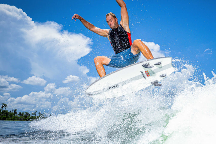 A man surfing on the 2025 Phase 5 Ahi Stealth surfboard, mid-air above the water with spray and a bright blue sky background.