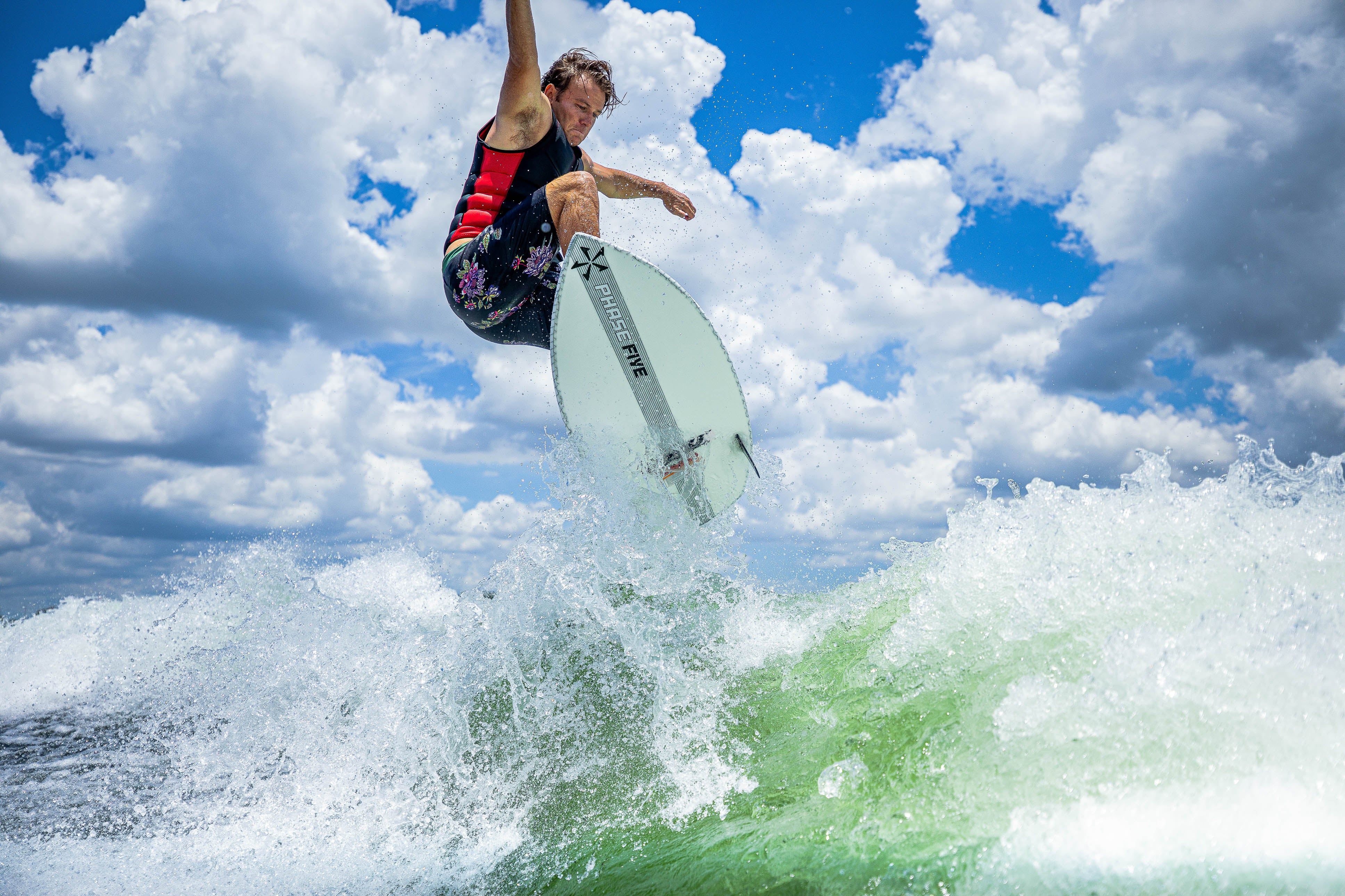 A surfer performs an aerial trick on a white Aku Surf Board with a blue sky and clouds in the background.