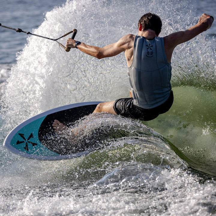 A person riding the 2026 Phase Five Avenger wakesurf board, carving through a wave with spray flying around.