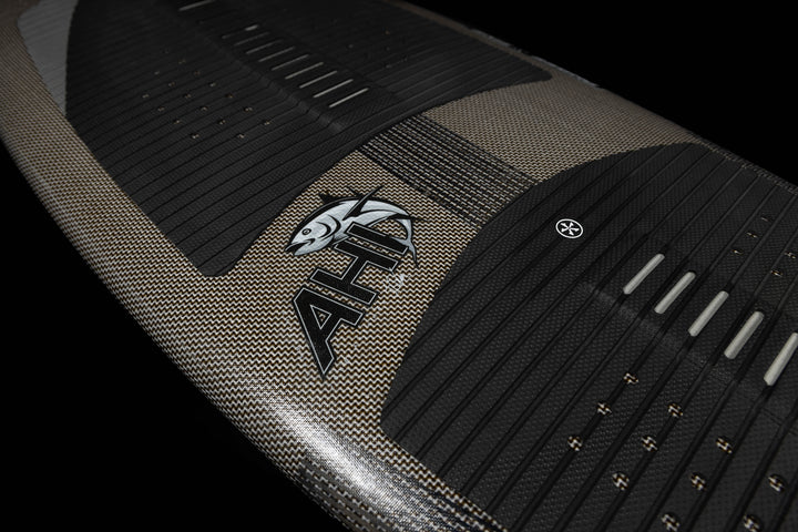 Close-up of the 2025 Phase 5 Ahi Stealth surfboard featuring textured grip pads, a logo with a fish graphic, and sleek black traction pads.