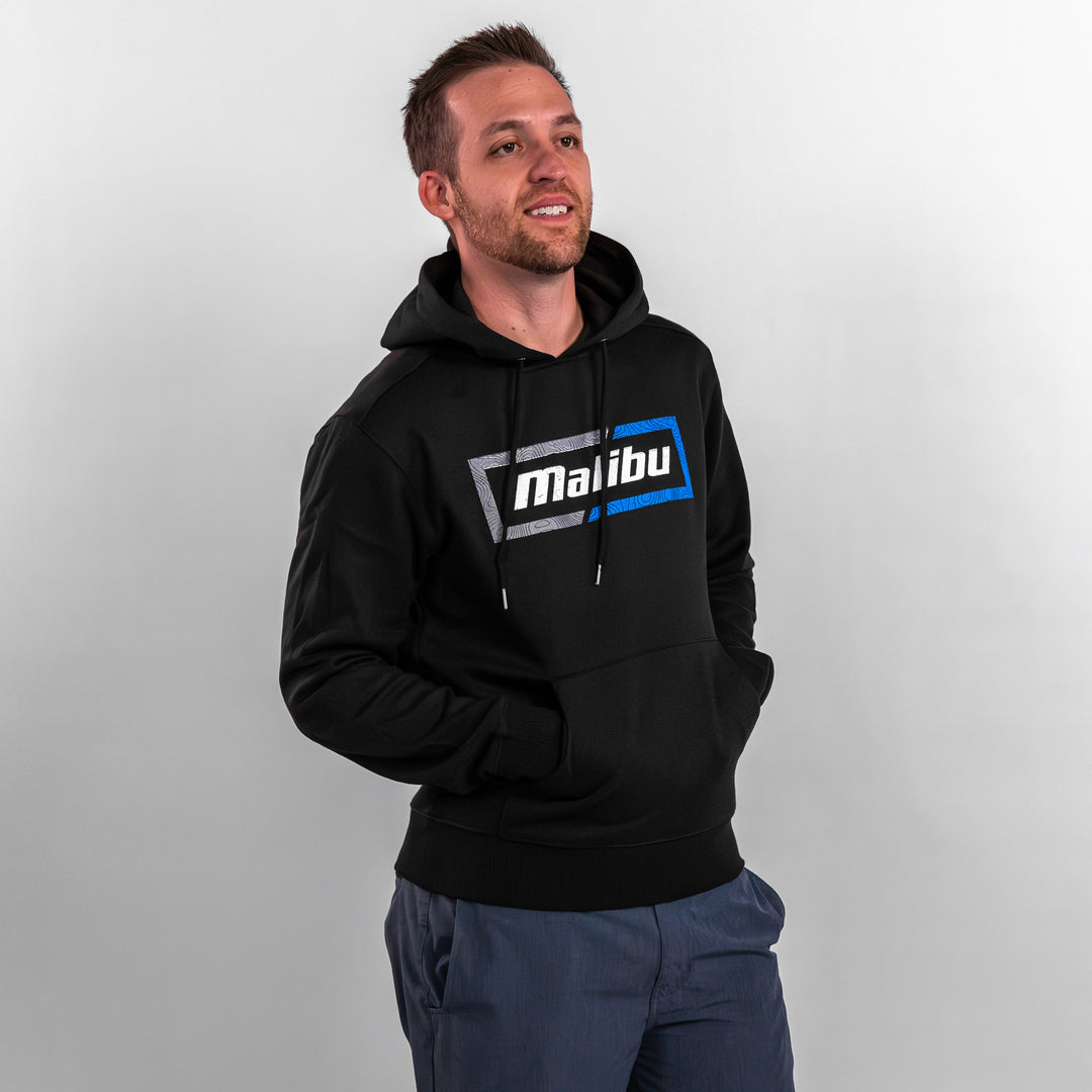 Man wearing a black Topo Hoodie featuring the Malibu logo in blue and gray, standing with hands in pockets.