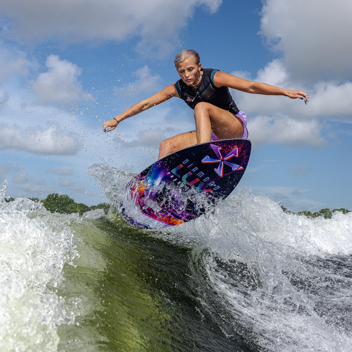 A person performs a wake surf maneuver on a colorful Phase Five Diamond Luv board, creating a splash in the water.