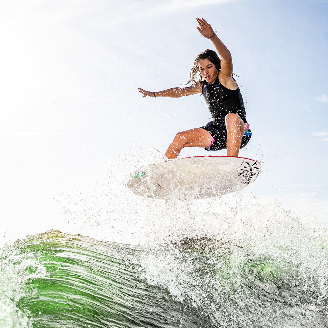 A young woman performs a jump on a 2025 Phase 5 Fish Skim Board over a wave, showcasing skill and excitement in water sports.
