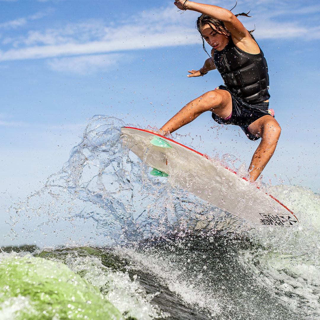 A young person performs a jump on a 2025 Phase 5 Fish Skim Board, splashing water in a dynamic action shot.