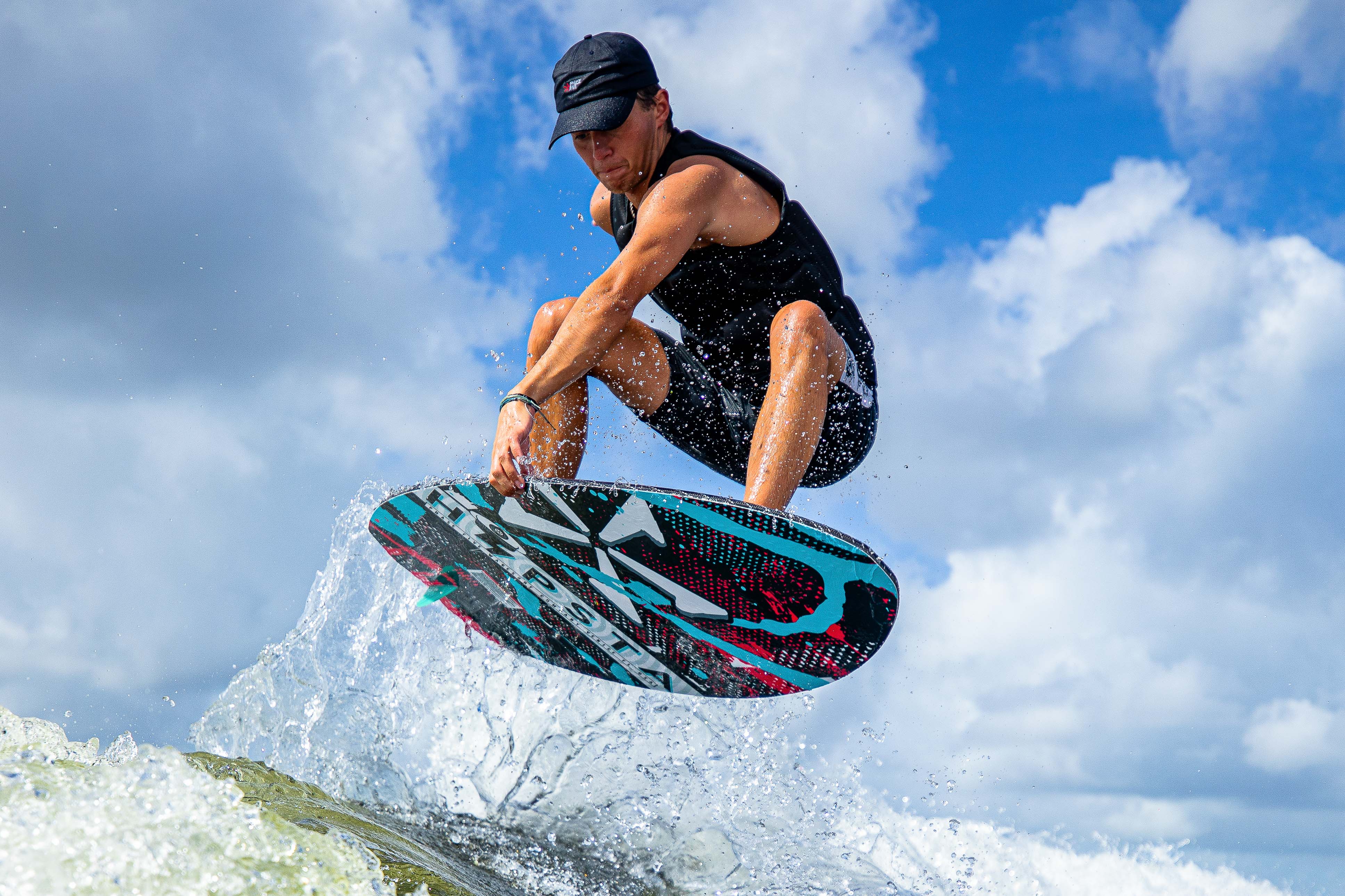 A person performs a jump on a colorful 2025 Phase 5 Hypsta skim board, splashing water against a cloudy sky backdrop.