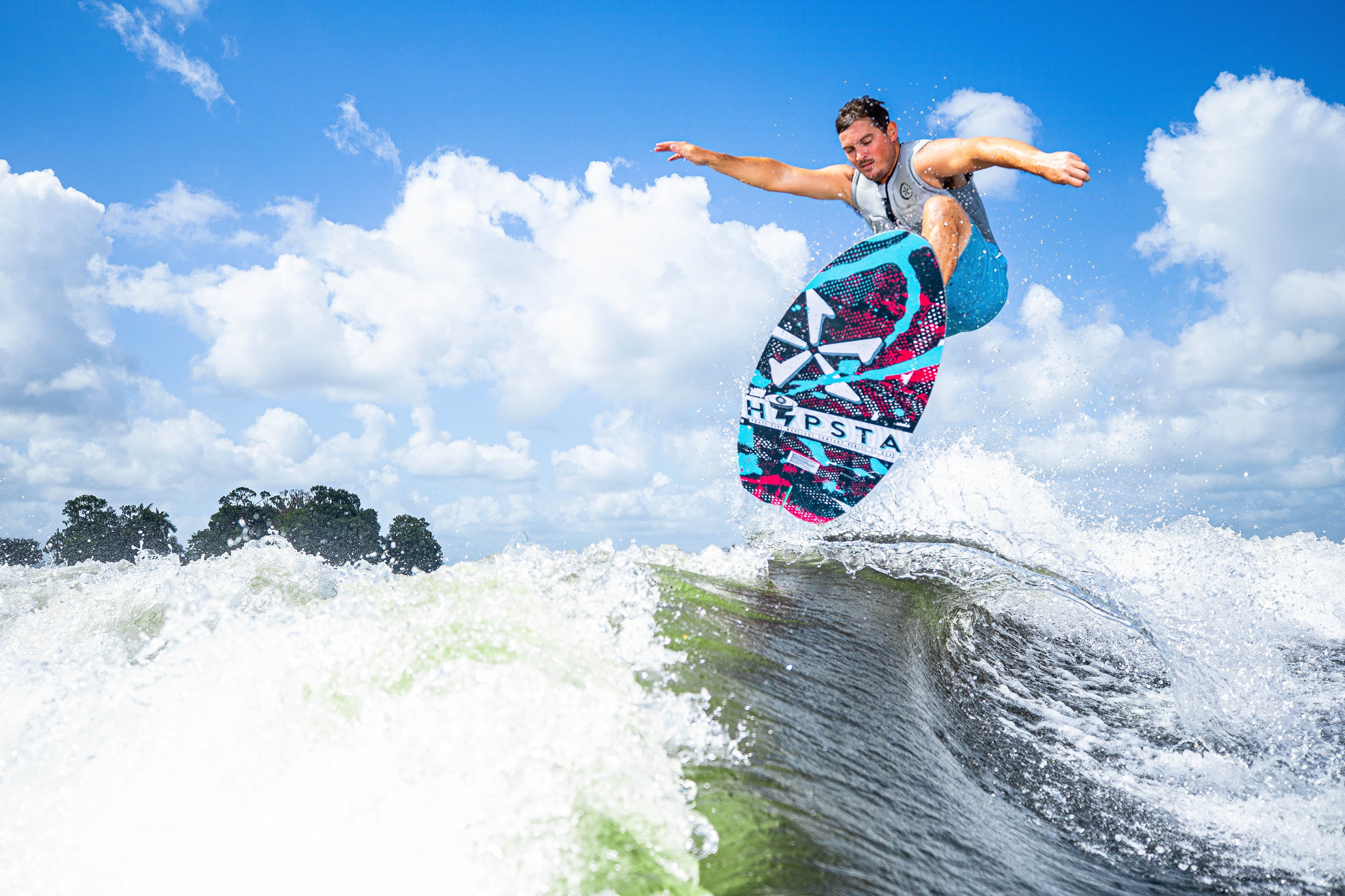A person performs a jump on a 2025 Phase 5 Hypsta skim board over a wave, showcasing vibrant colors and a clear sky.