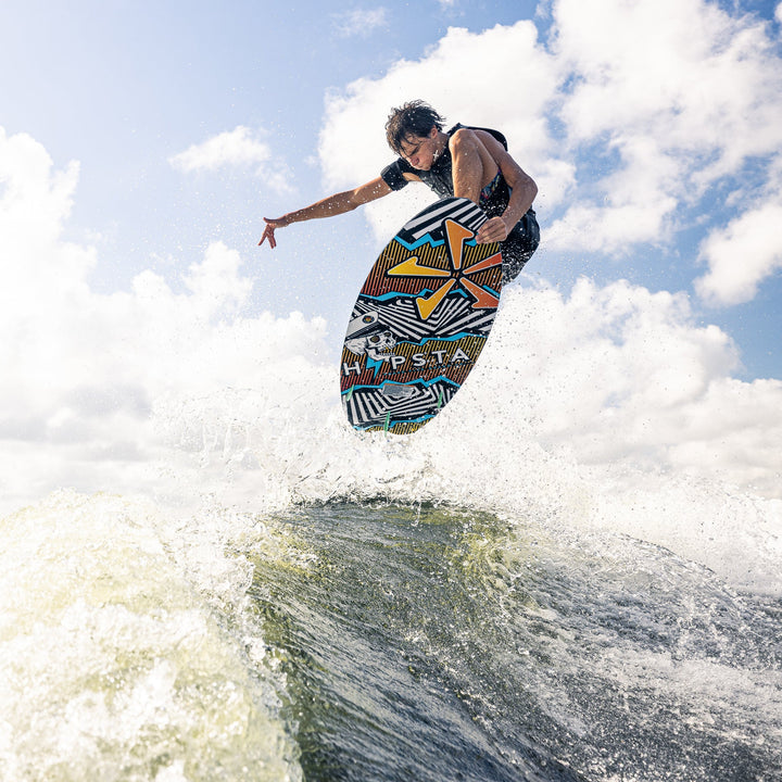 A rider performs a jump on the 2026 Phase Five Hypsta V2 wake surfboard, showcasing its vibrant design against a cloudy sky.